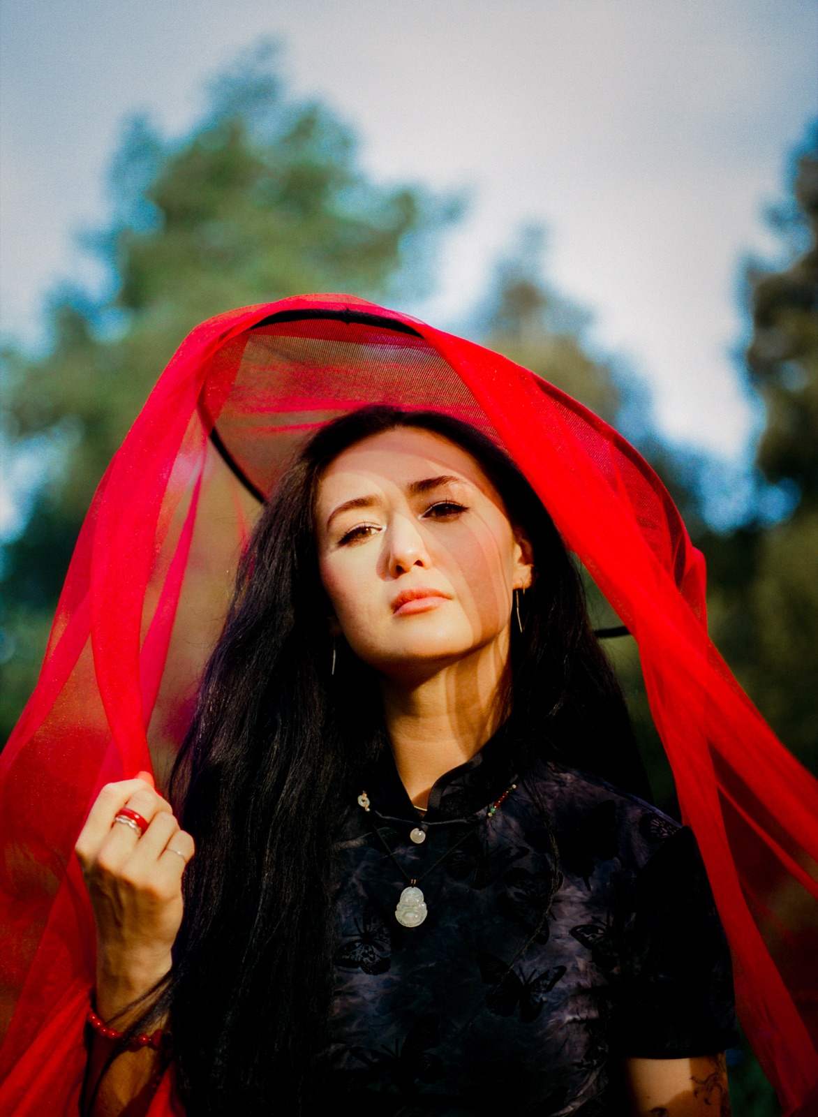 Portrait of dān dān beneath a red tulle veil held above her head, long dark hair loose, wearing a black butterfly-print top with a jade pendant, trees soft in the background.