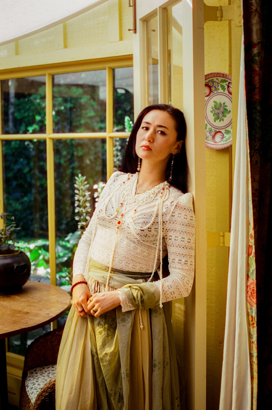 dān dān leans against a yellow window frame in a sunroom in a white lace top and pearls
