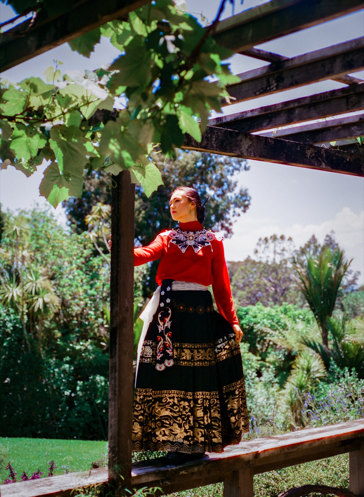 dān dān stands on a garden pergola bench in a red top and embroidered black and gold skirt