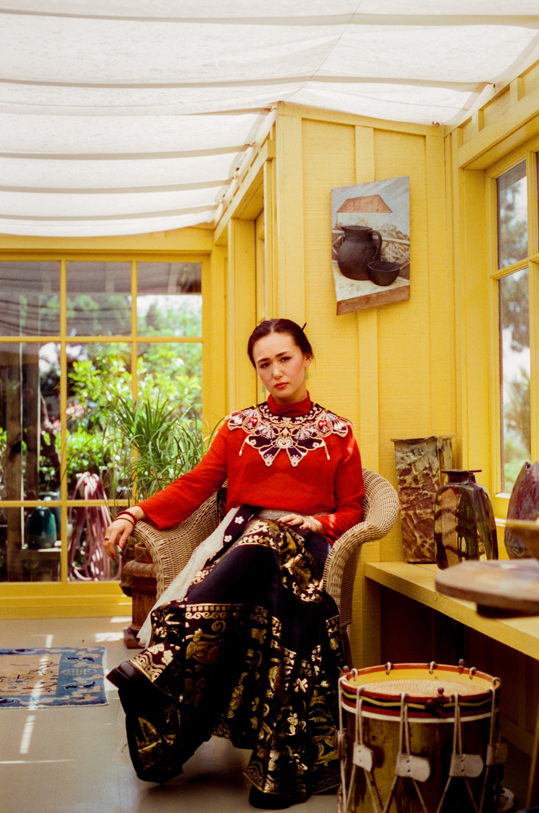 dān dān sits in a wicker chair in a yellow sunroom
