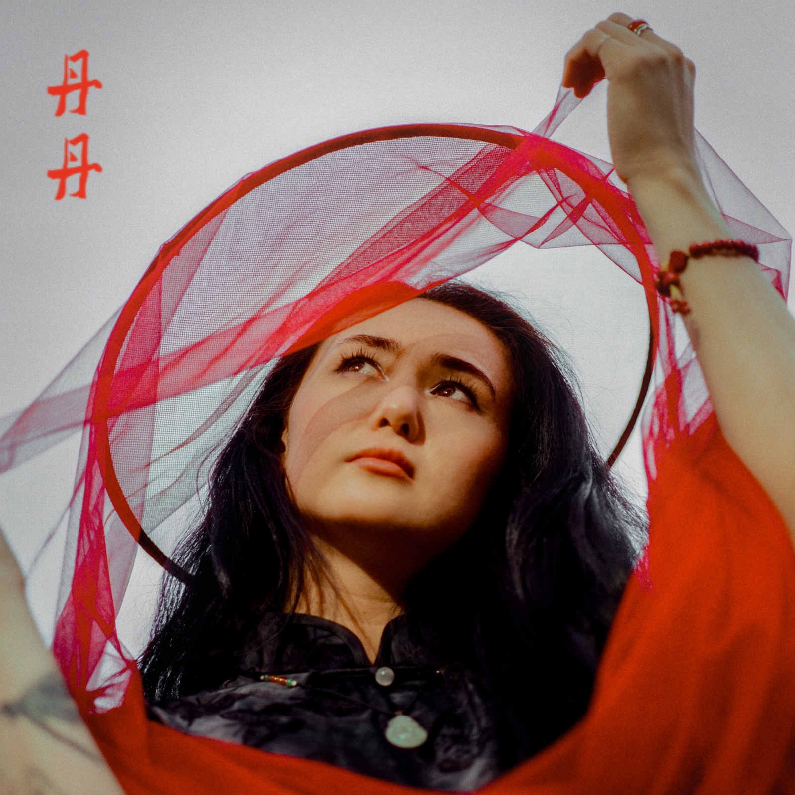 dān dān looks upward through a ring of red tulle held above her head, wearing a red and black outfit with a jade pendant, against a pale sky. Cover photography by Flora Xie.